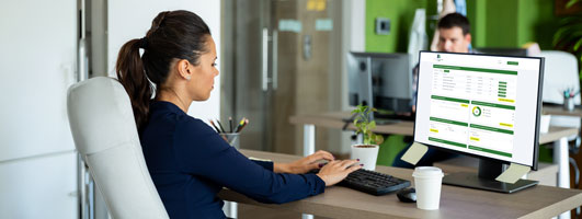 Mujer teclea frente a un monitor con un panel de indicadores, con un hombre trabajando al fondo